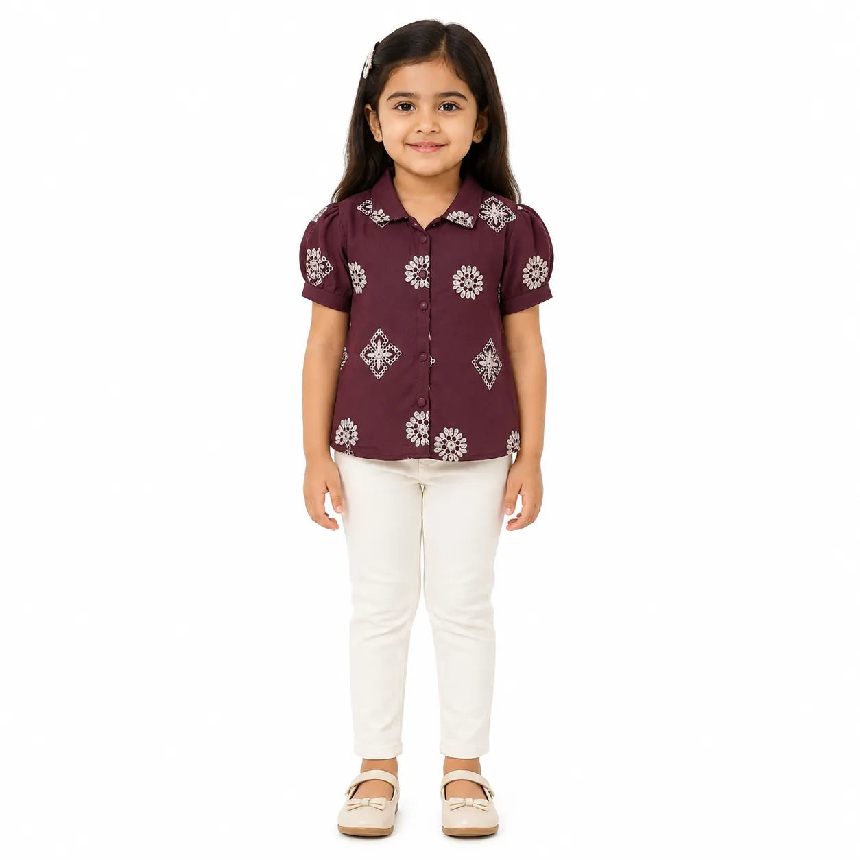A young girl standing against a white background, wearing a short-sleeve maroon button-up shirt with white floral patterns, paired with off-white pants and beige shoes.