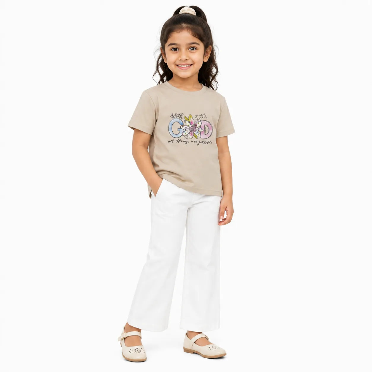 A young girl stands against a plain white background wearing a beige graphic T-shirt, white wide-leg pants, and beige flats. The T-shirt has a colorful bicycle graphic with the words 'AMG GOOD' and 'stay good to yourself.