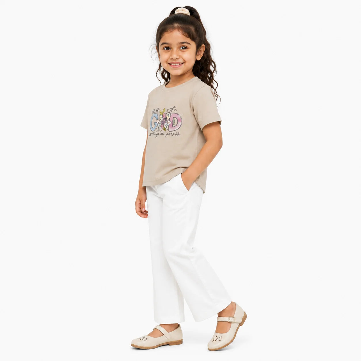 A young girl with her hair in a ponytail stands in a studio wearing a beige graphic t-shirt, white wide-leg pants, and beige flat shoes. The background is a clean, solid white.