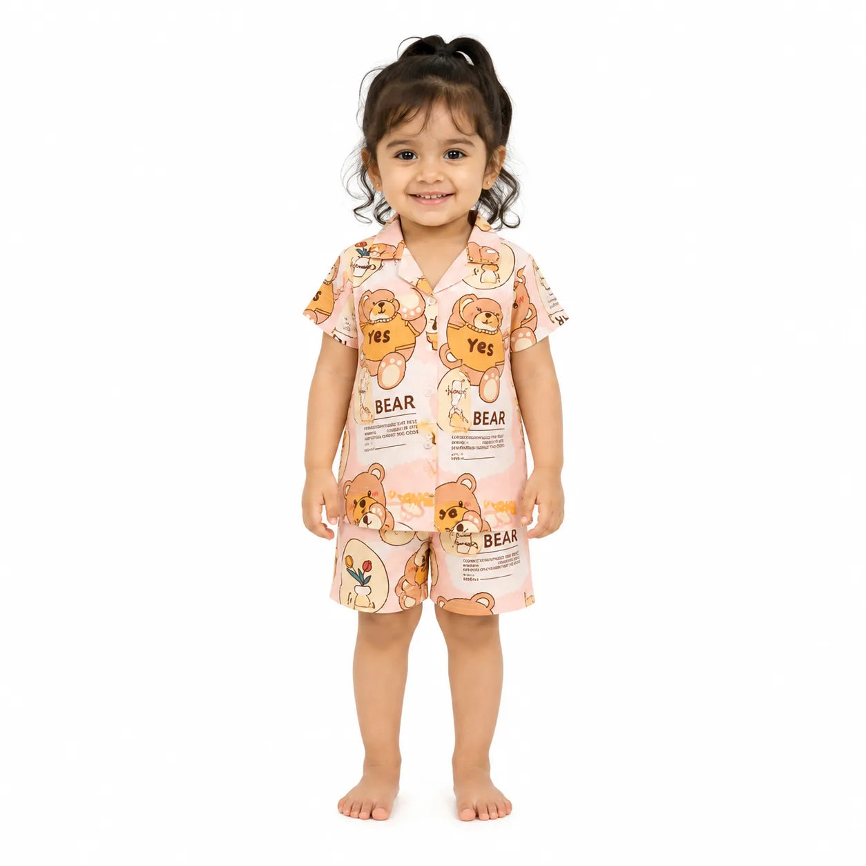 A toddler girl stands barefoot against a plain white background wearing a two-piece pajama set. The pajama set consists of a short-sleeve top and matching shorts in a light pink color, both printed with a cute cartoon bear pattern in orange, brown, and white, along with the word 'BEAR'.